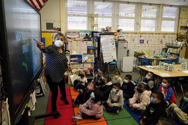 An adult stands in front of a group of children, pointing at something on a projected screen