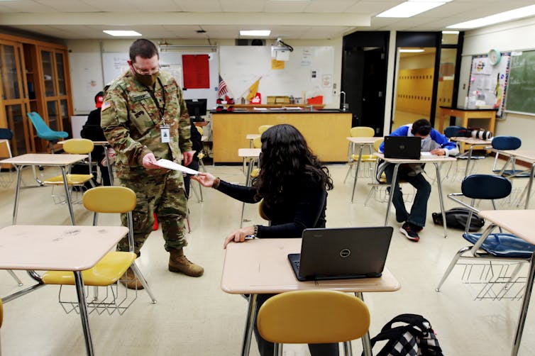 A man in camouflage stands in a classroom and hands a piece of paper to a student