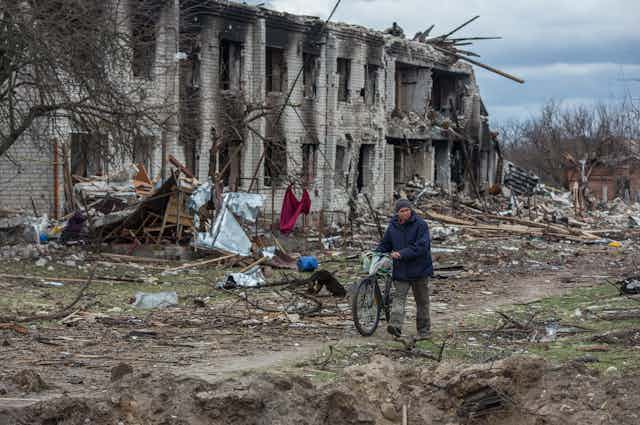 Un hombre camina al lado de su bicicleta por un descampado al lado de un edificio bombardeado.