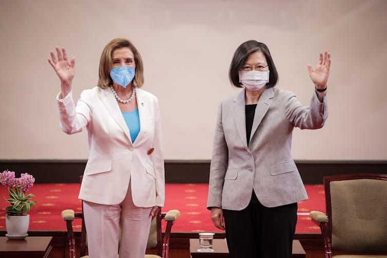 A white woman in a beige pantsuit and blue face mask stands next to a middle-aged Asian woman also wearing a pantsuit. Both wave their hands.