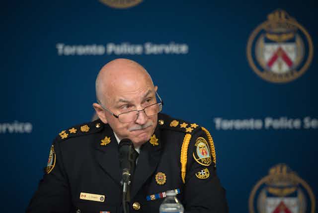 A bald man wearing a police uniform sits in front of a microphone