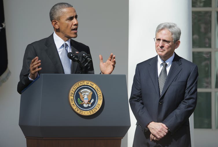 A man at a lectern in a suit and tie gestures with his hands while another man, also in a suit and tie, stands next to him, listening.