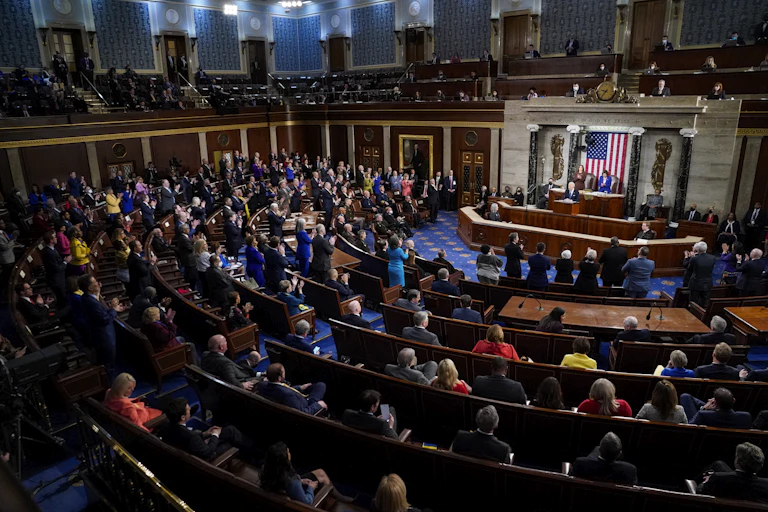 A man in a large chamber speaking to an audience in front of an American flag.