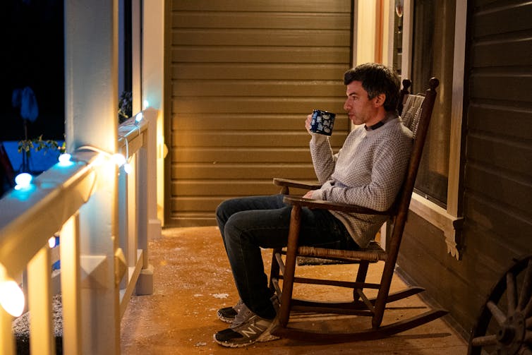 Nathan Fielder sits in a rocking chair on a porch.
