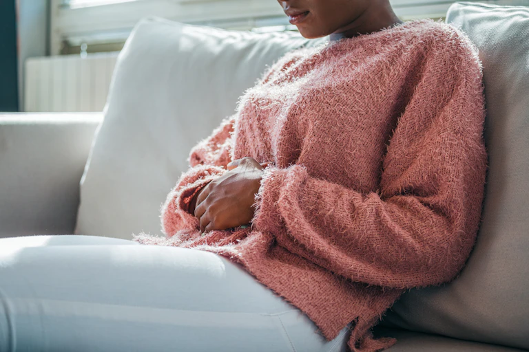 African-Australian woman in a pink jumper holds her abdomen, in pain