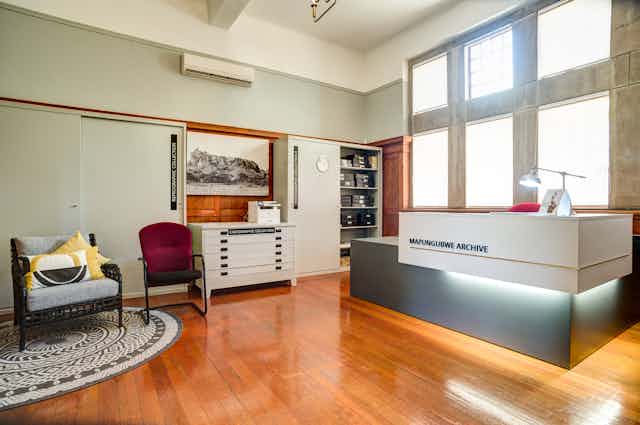 A room with wooden floors and a modern, angular reception desk with lettering 'Mapungubwe Archive' and chairs and cabinets containing archival material.