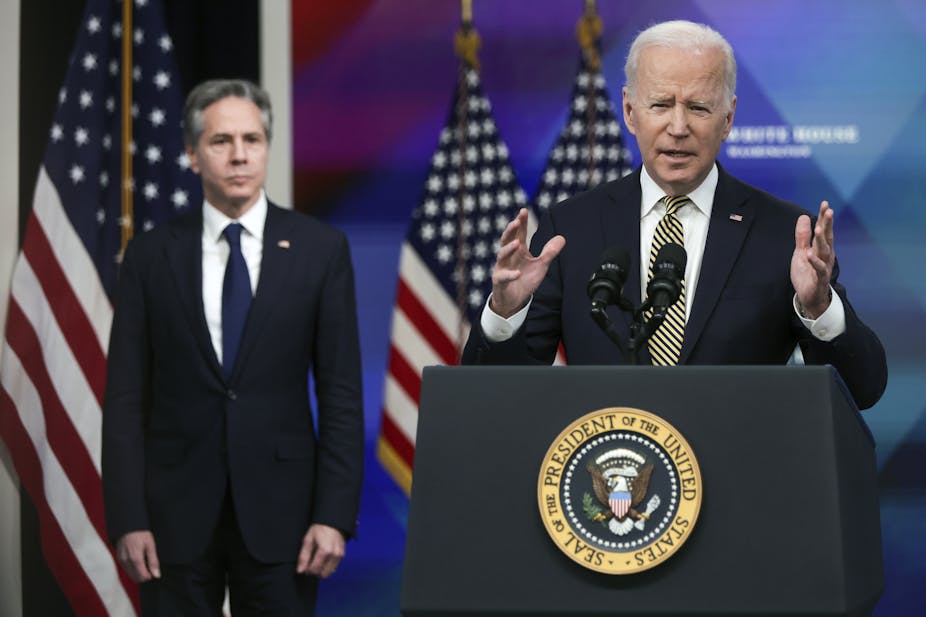 One man speaks at a podium carrying the seal of the United States, another man stands in the background in front of American flags