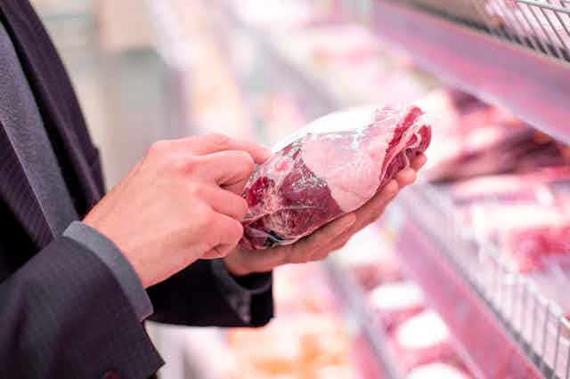 A man inspects a plastic-wrapped joint of meat in a supermarket aisle.