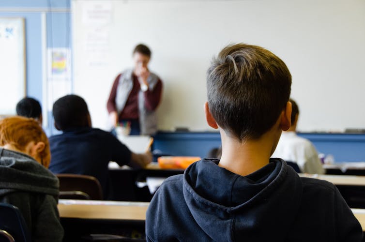 Desde la parte de atrás de la clase, la fotografía se enfoca en un niño que atiende atentamente las explicaciones del profesor en la pizarra