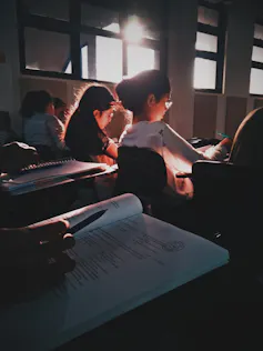 En primer término, un cuaderno de notas en el pupitre de una clase en la que los alumnos escriben (probablemente un examen).