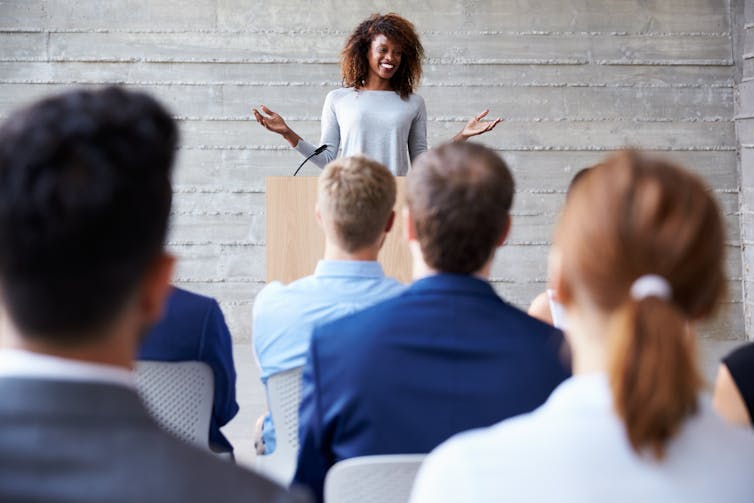 A smiling woman stands and speaks at a podium while an audience looks on.