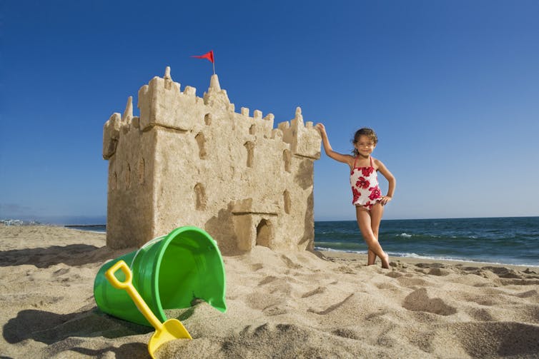 On a sandy beach, with a plastic green bucket and yellow shovel in the foreground, a blue sky and bluer water in the background, a colorfully swimsuited girl stands with one hand on her hip and the other on a sand castle as tall as she is.