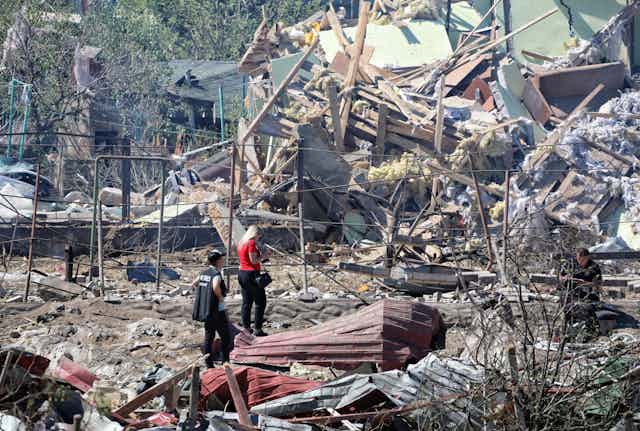 Two people are at a distance standing amidst rubble and a collapsed building.