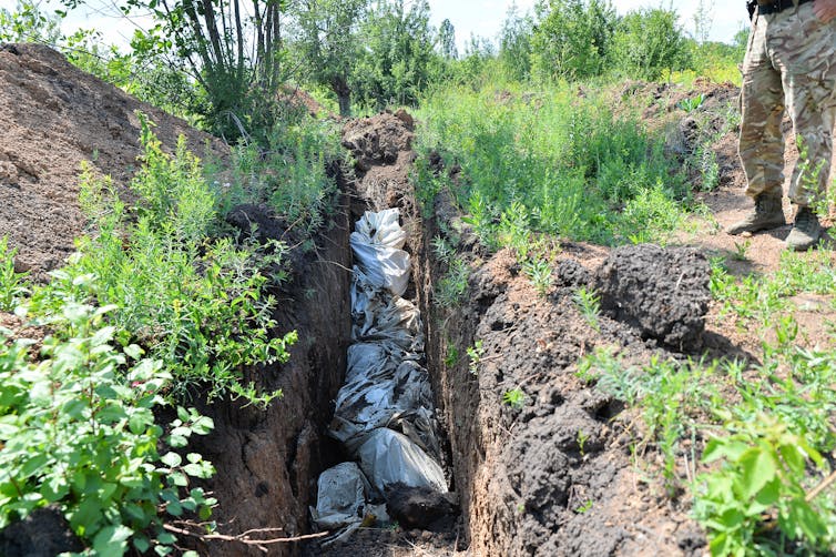 A trench in the ground shows stuffed white garbage bags lined up. One person is shown from the waist down observing the bags and the trench.