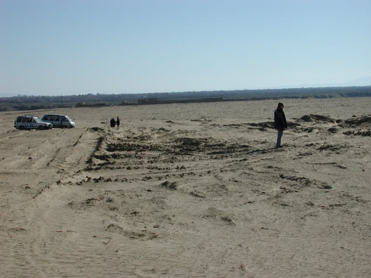 A person stands in a dirt field with 2 U.N. trucks in the background.
