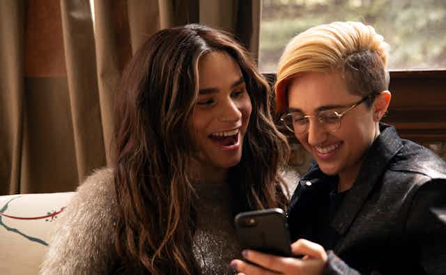 A transfeminine non-binary person and transmasculine gender-nonconforming person looking at a phone and laughing.