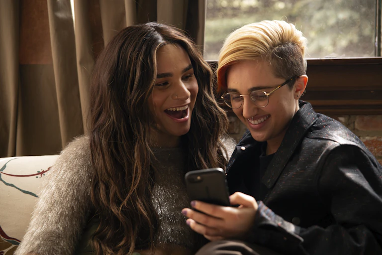 A transfeminine non-binary person and transmasculine gender-nonconforming person looking at a phone and laughing.