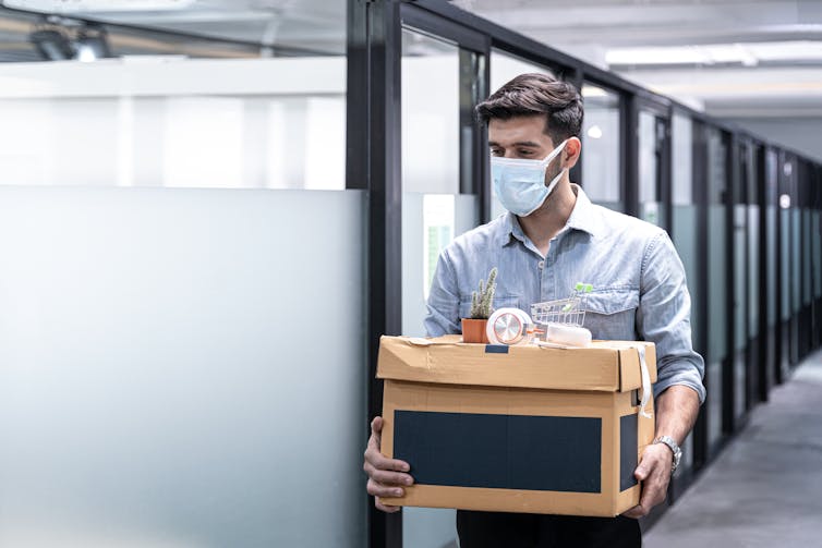 A man wearing a mask carrying a box of belongings past a row of cubicles