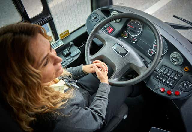 A bus driver sitting behind a steering wheel with her hands folded on her lap