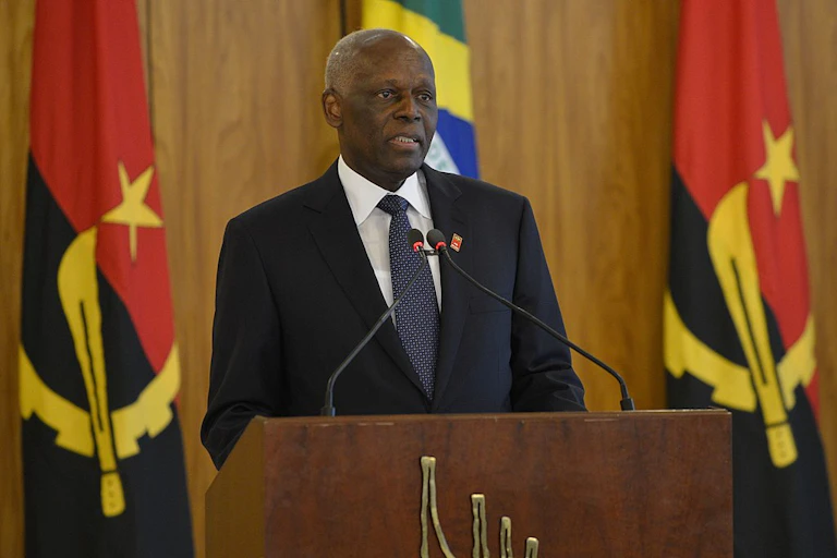 A bald-headed man wearing a suit and tie speaks into two small microphones at a podium, with his back to two Angolan flags showing a hammer and circle on either side.