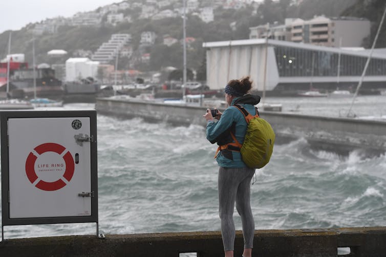 Woman taking photos of huge waves hitting the coastline of downtown Wellington.
