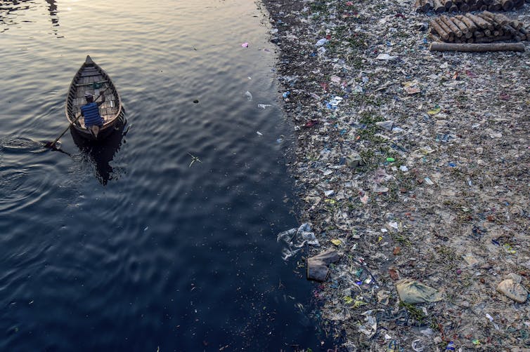 Viewed from above, a man paddles a wide canoe down a river laden with plastic and other waste.