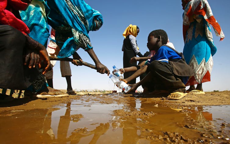 A boy crouches next to a puddle where a woman is filling plastic water bottles with a hose.