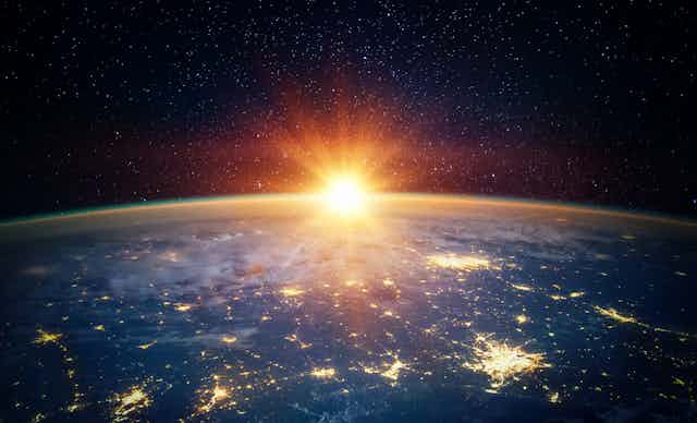 The Earth is shown at an angle from space, with cities lit up down below, and the Sun partially creeping up from the horizon. 