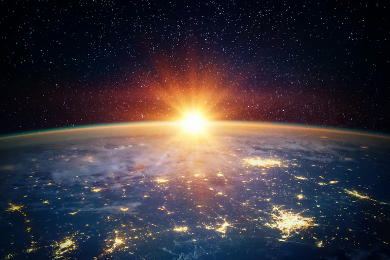 The Earth is shown at an angle from space, with cities lit up down below, and the Sun partially creeping up from the horizon.