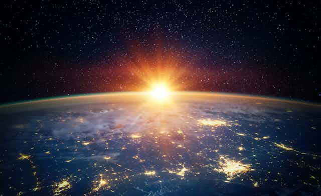 Tierra vista desde el espacio, con las ciudades iluminadas abajo, y el Sol asomando parcialmente desde el horizonte. 