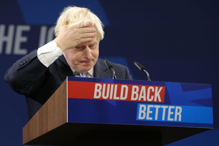 Boris Johnson clutches his forehead during a speech behind a podium branded with the slogan 'build back better'.