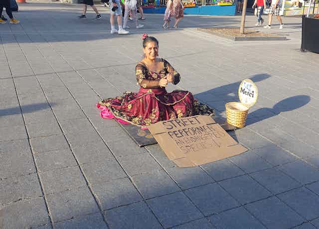 A woman sits on the street wearing a red dress, in front of her a sign reads 'street performer an endangered species'