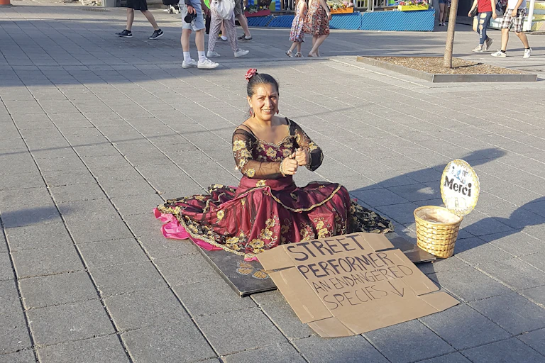A woman sits on the street wearing a red dress, in front of her a sign reads 'street performer an endangered species'