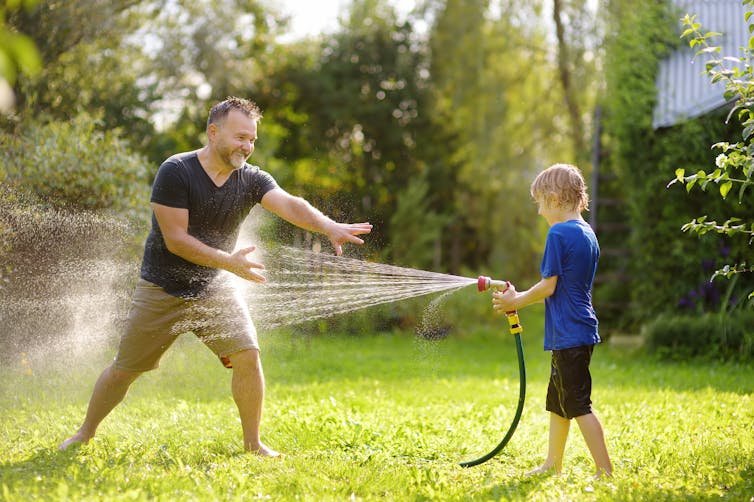 A young boy playfully sprays his father with a hosepipe in a lush, green garden.