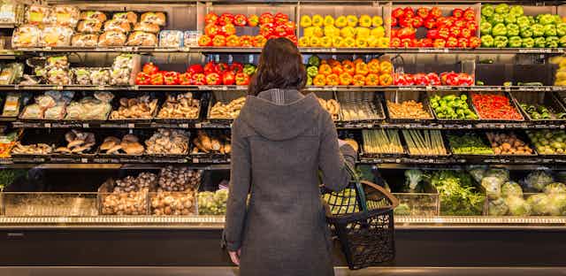 A shopper with a shopping basket over their arm facing a shelf full of groceries.