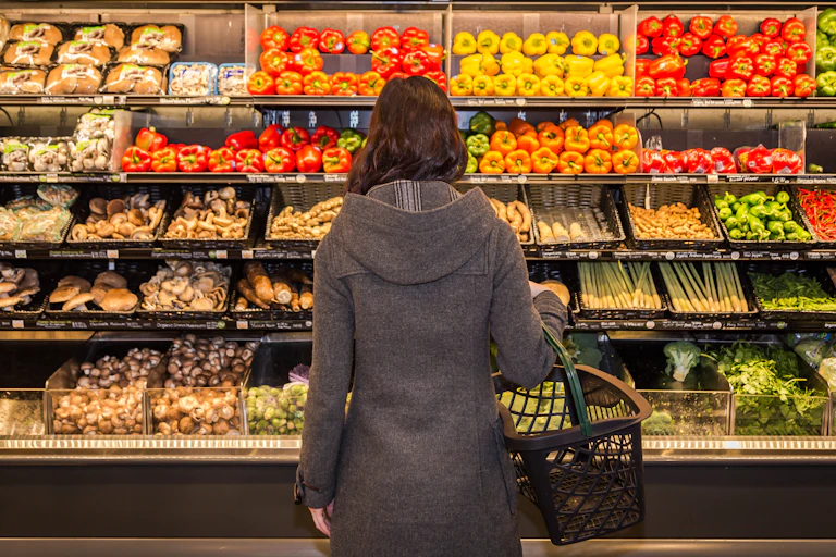 A shopper with a shopping basket over their arm facing a shelf full of groceries.