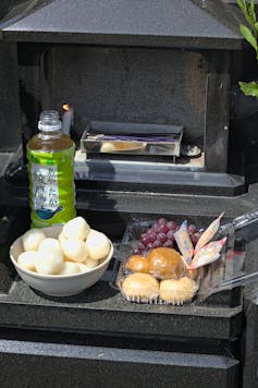 A plastic bottle of tea and a bowl of rice cakes with other treats in a plastic container on a grave stone.
