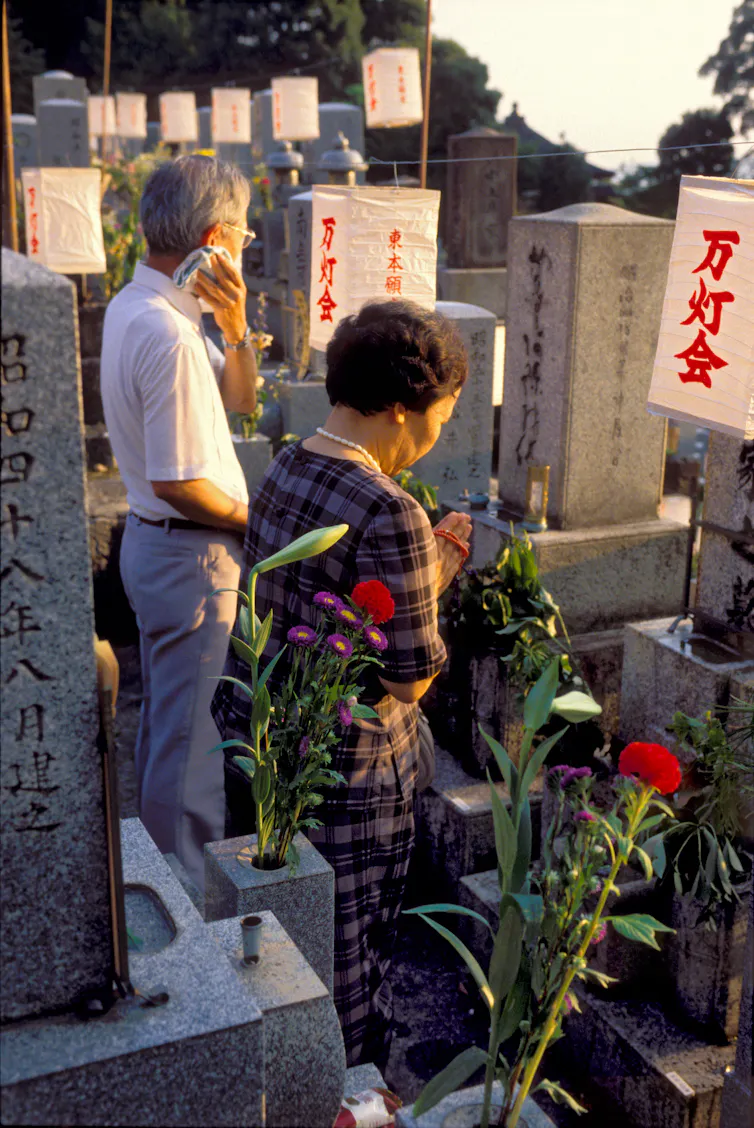 Two people in front of grey grave stones with lanterns in the sunlight.