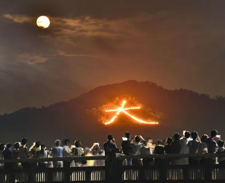 A crowd looks on at a bonfire in the shape of a Japanese character on a hillside under the moonlight.