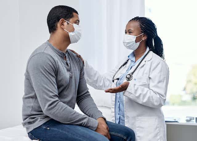 A woman in a mask and white coat with a stethoscope talking to a man with her hand on his shoulder