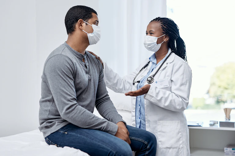 A woman in a mask and white coat with a stethoscope talking to a man with her hand on his shoulder