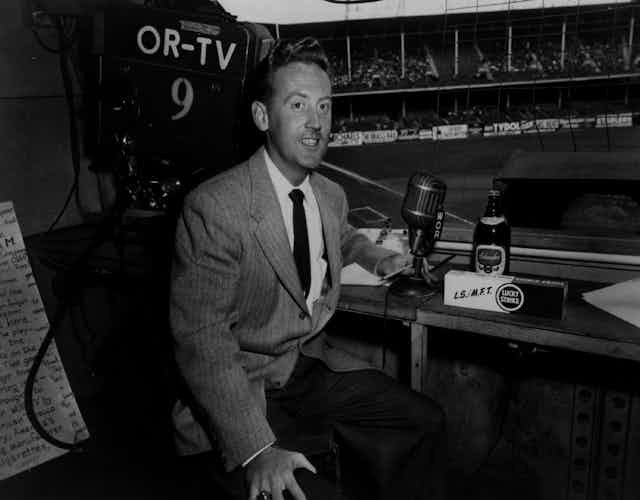 Man in suit sits before microphone at baseball stadium.