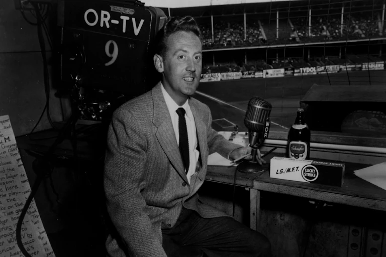 Man in suit sits before microphone at baseball stadium.