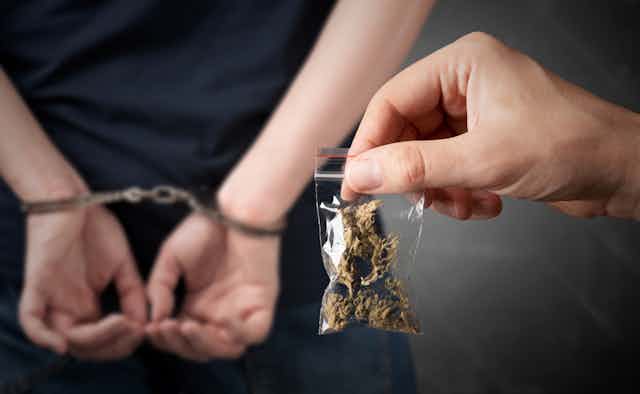 A hand holding up a small bag of cannabis in front of someone with handcuffs on.