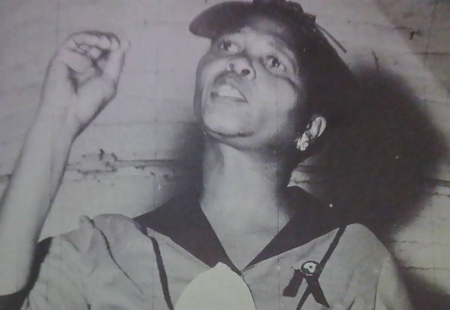 A woman holds up a hand to make a point as she speaks earnestly, a cap on and wearing a uniform.
