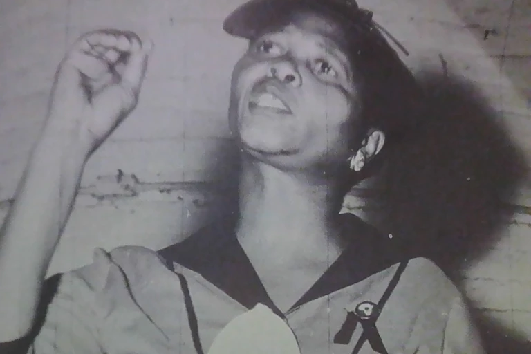 A woman holds up a hand to make a point as she speaks earnestly, a cap on and wearing a uniform.
