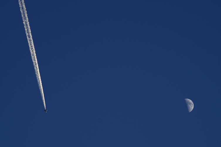 A contrail from a military jet in a blue sky with a half-moon visible.