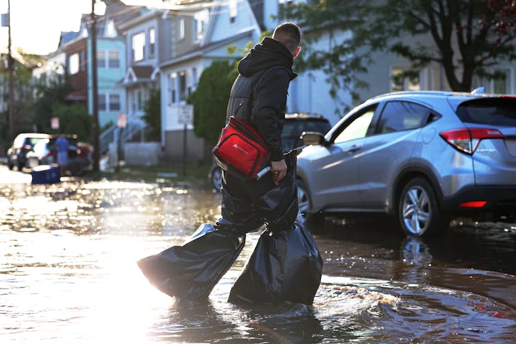 A person wearing trash bags around each leg to keep them dry crosses a flooded city street in New Jersey. An overturned trash can floats in the background.