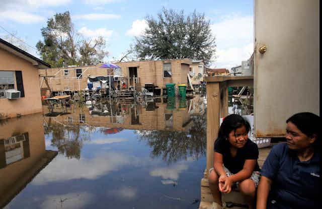 A woman and child sit on the steps of a home above a flooded and hurricane-damaged neighborhood.