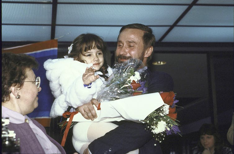 A man in a suit smiles and holds a young girl in a white jacket, who waves at the camera.